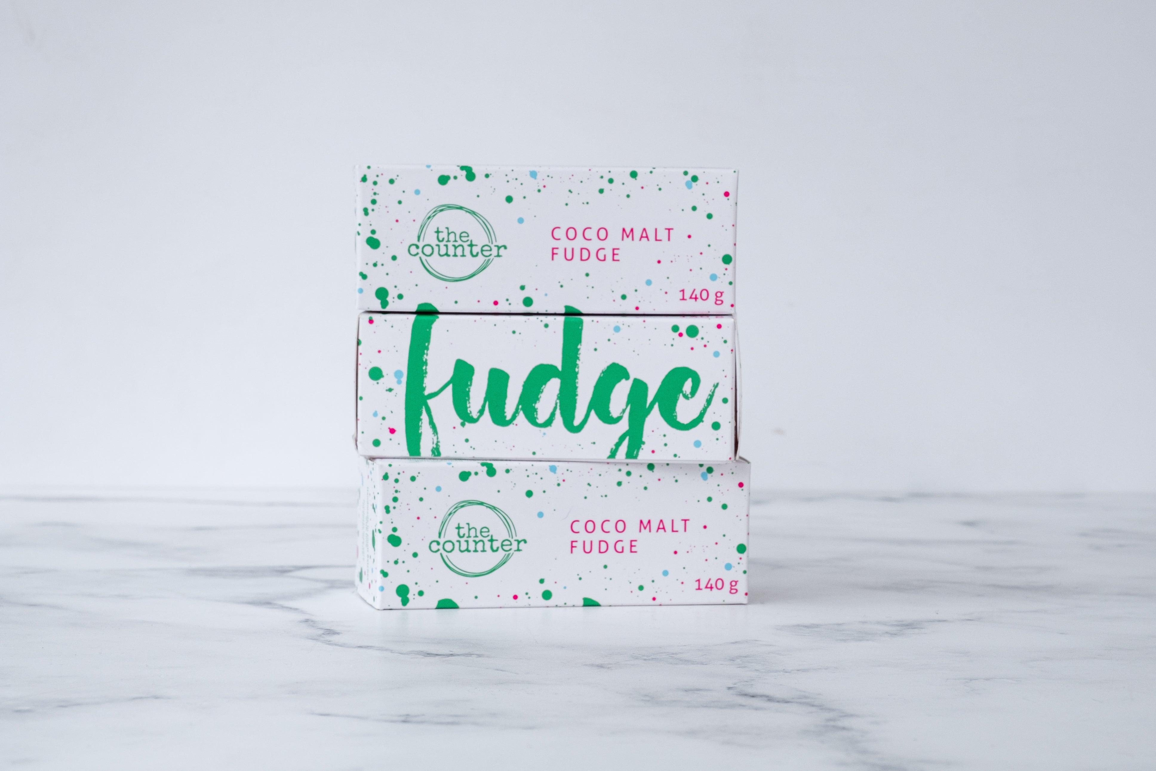Three white fudge boxes are stacked on a neutral background. Paint splashes in hunter green and pink surround a messy circled logo for The Counter and the words 'Coco Malt fudge' are written in cerise pink block lettering on the first and third box. The middle box has large hunter green lettering states 'fudge' and is surrounded by hunter green and cerise pink paint splashes.