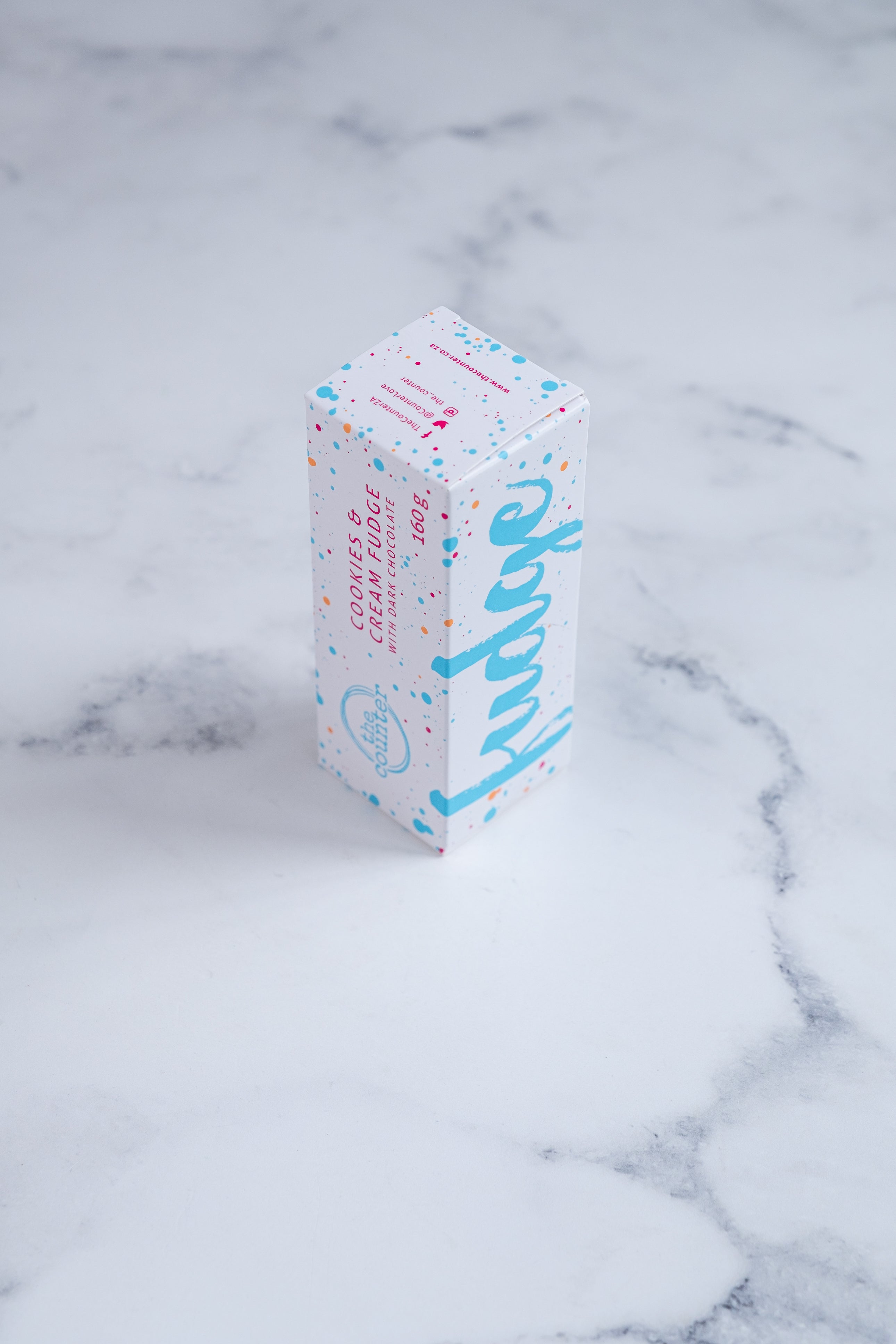 A white box stands upright on a neutral background. Large bright blue lettering states 'fudge' and is surrounded by bright blue and cerise pink paint splashes. A messy circled logo for The Counter and the words 'Cookies & Cream fudge with dark chocolate' are written in cerise pink block lettering.