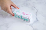 Load image into Gallery viewer, A hand with red nail polish grabs a White chocolate fudge box with &#39;The Counter&#39; logo in turquoise, surrounded by several thin lines creating a circle. Pink text stating &#39;white chocolate fudge&#39; and paint splashes surrounding the text and logo.

