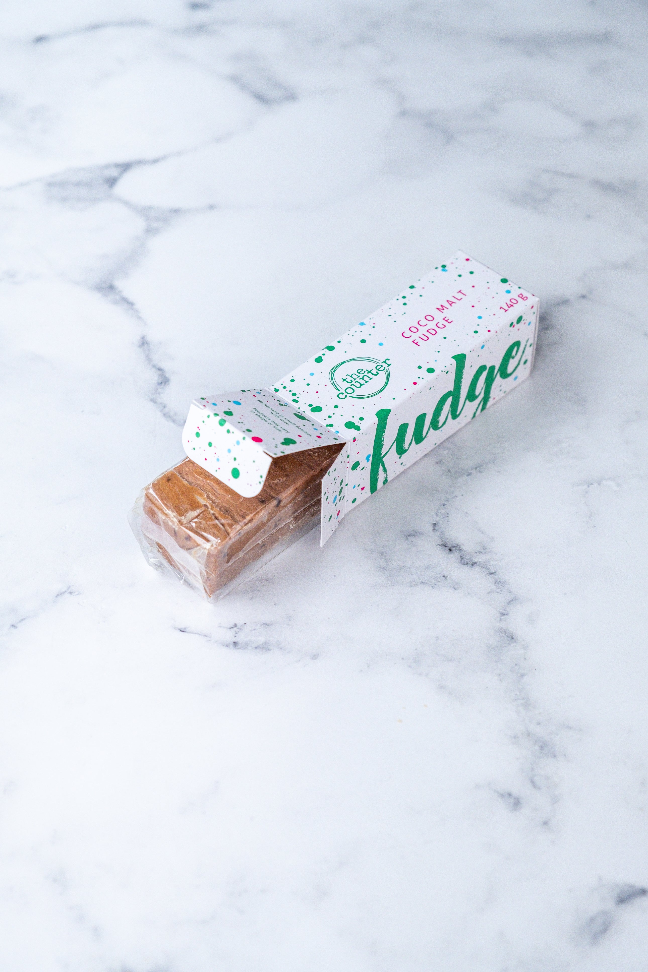 A large bar of Coco Malt (Milo) fudge peeks our from a white box. Paint splashes in hunter green and pink surround a messy circled logo for The Counter and the words 'Coco Malt fudge' are written in cerise pink block lettering.