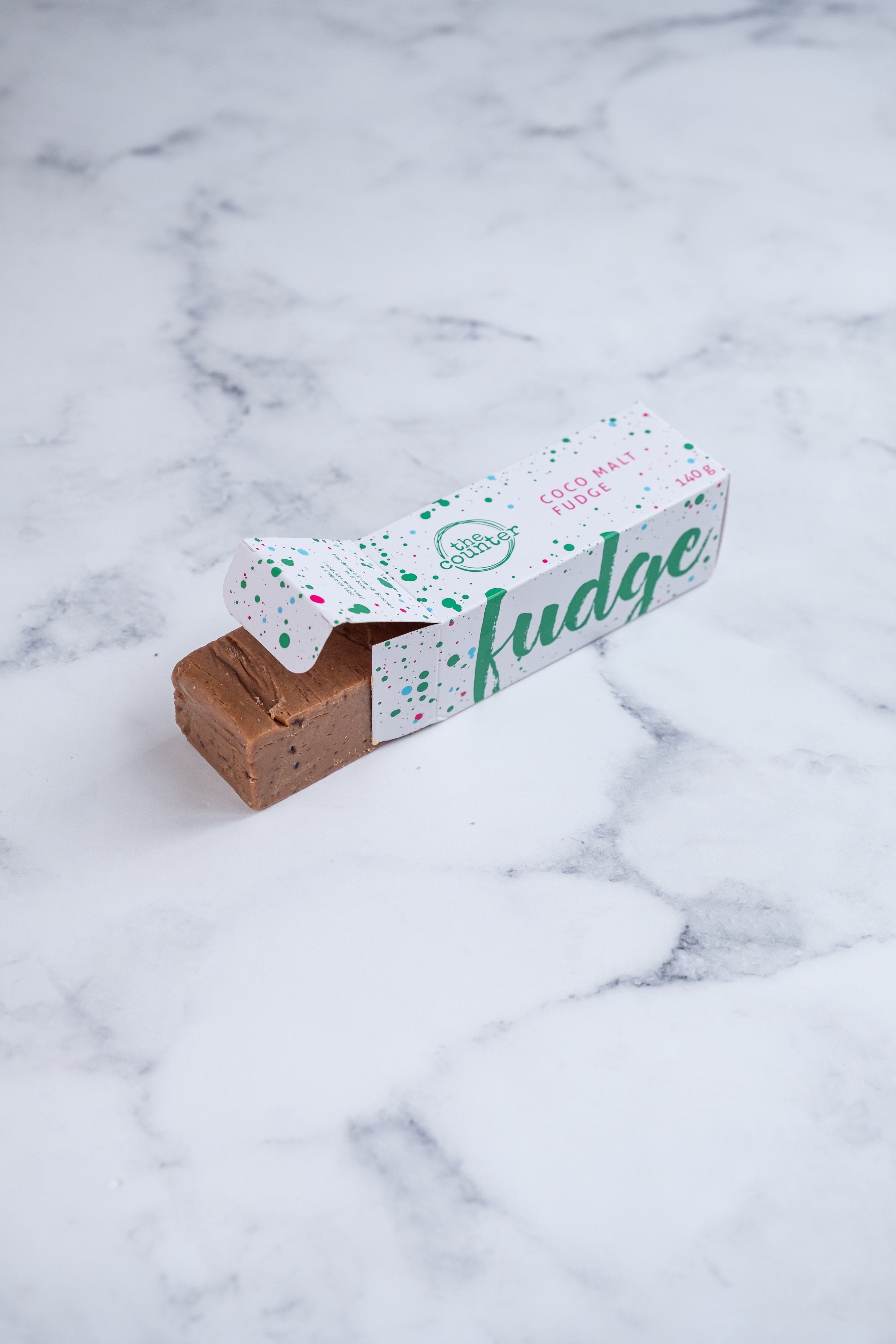 A large bar of Coco Malt (Milo) fudge peeks our from a white box. Paint splashes in hunter green and pink surround a messy circled logo for The Counter and the words 'Coco Malt fudge' are written in cerise pink block lettering.