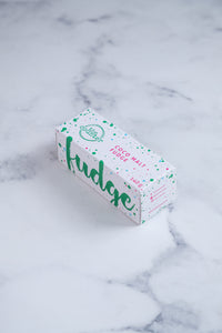 A white box sits on a neutral background. Large hunter green lettering states 'fudge' and is surrounded by hunter green and cerise pink paint splashes. The other side of the box that is visible has Paint splashes in hunter green and pink surround a messy circled logo for The Counter and the words 'Coco Malt fudge' are written in cerise pink block lettering.