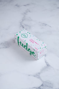 A white box sits on a neutral background. Large hunter green lettering states 'fudge' and is surrounded by hunter green and cerise pink paint splashes. The other side of the box that is visible has Paint splashes in hunter green and pink surround a messy circled logo for The Counter and the words 'Coco Malt fudge' are written in cerise pink block lettering.