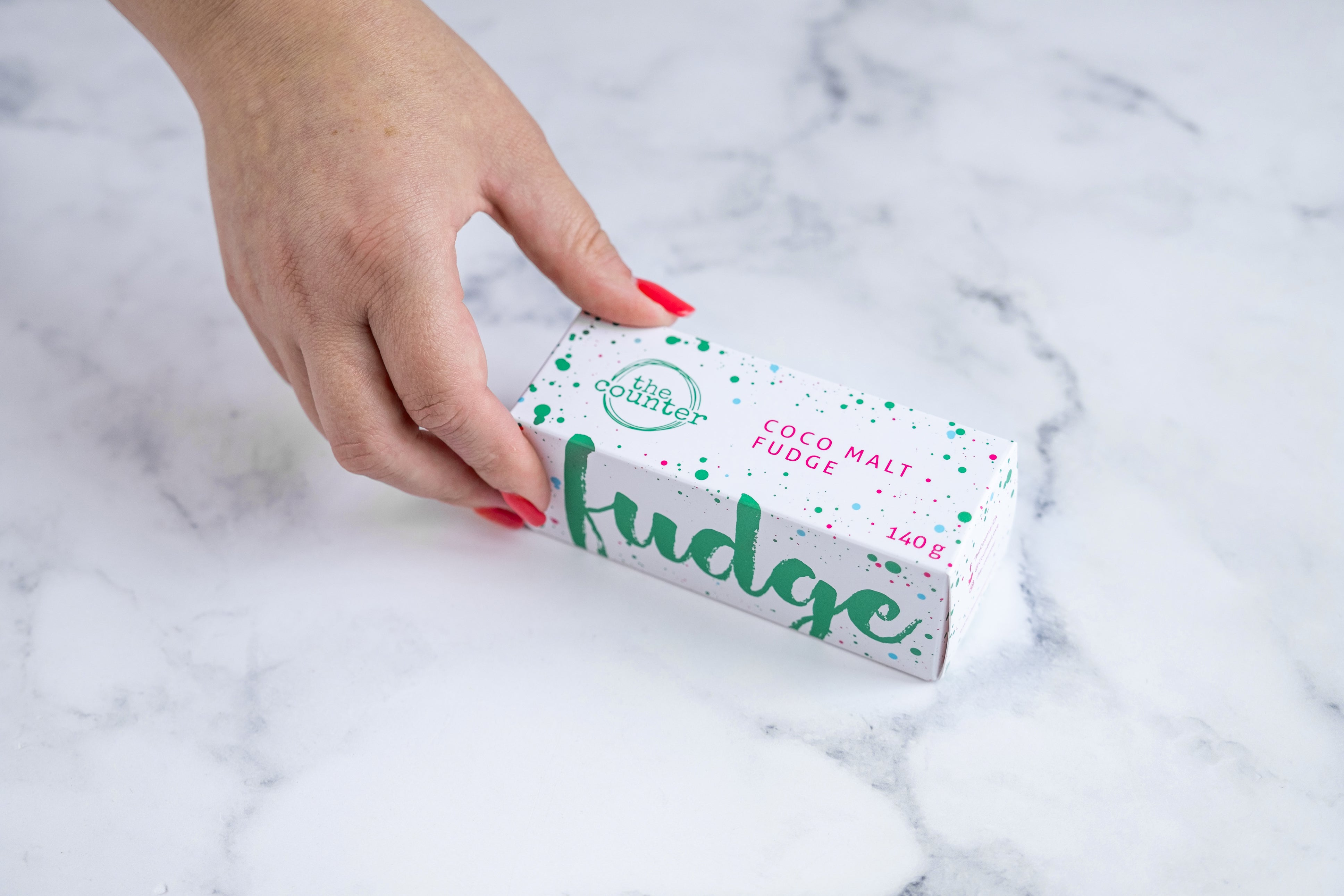 A white fudge box sits on a neutral background. Paint splashes in hunter green and pink surround a messy circled logo for The Counter and the words 'Coco Malt fudge' are written in cerise pink block lettering. On the topside of the box - large hunter green lettering states 'fudge' and is surrounded by hunter green and cerise pink paint splashes. A manicured hand with red nail polish grabs the box.