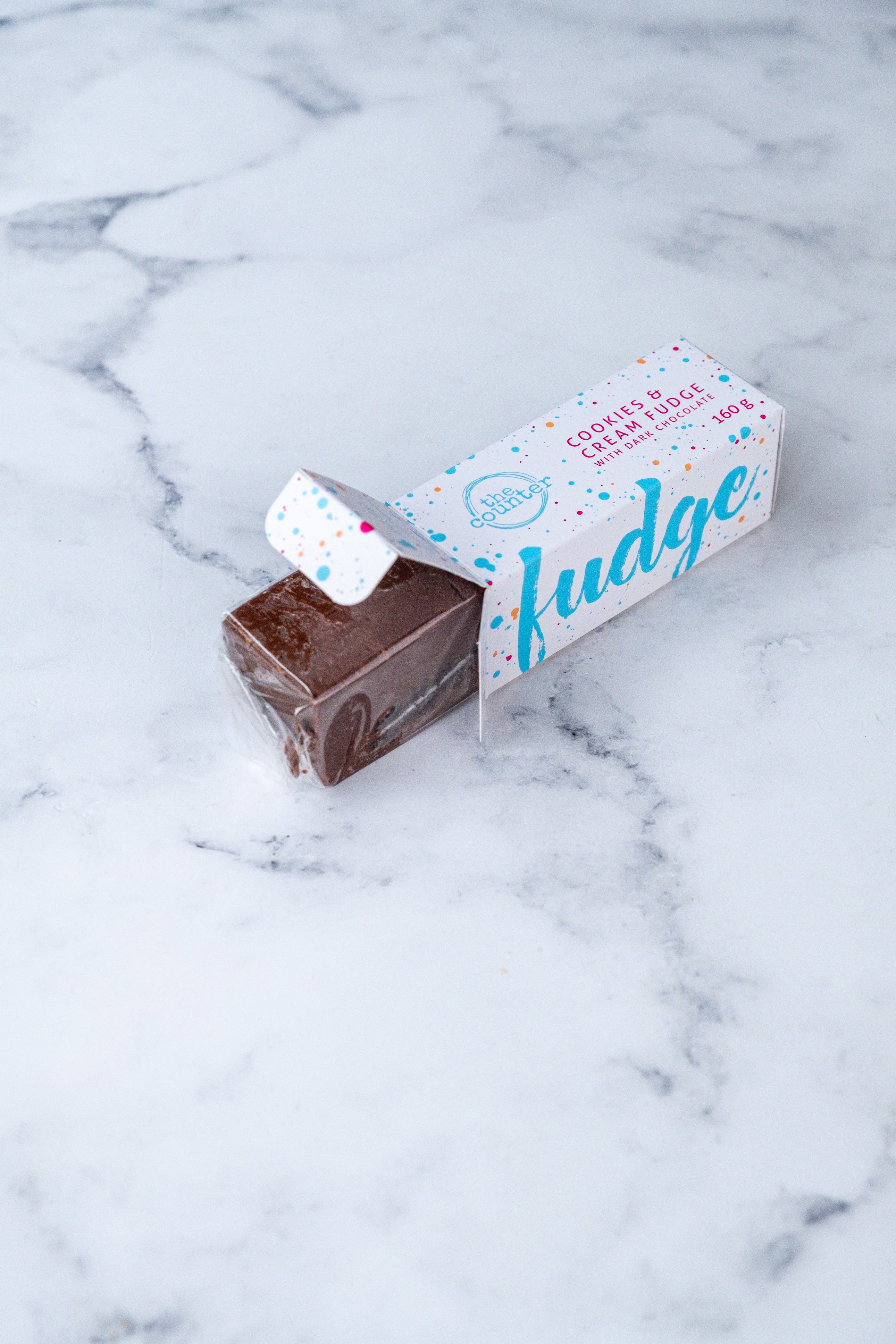 A large bar of dark chocolate and Oreo fudge (cookies and cream) peeks our from a white box. Paint splashes in bright blue and pink surround a messy circled logo for The Counter and the words 'Cookies & Cream fudge with dark chocolate' are written in cerise pink block lettering.