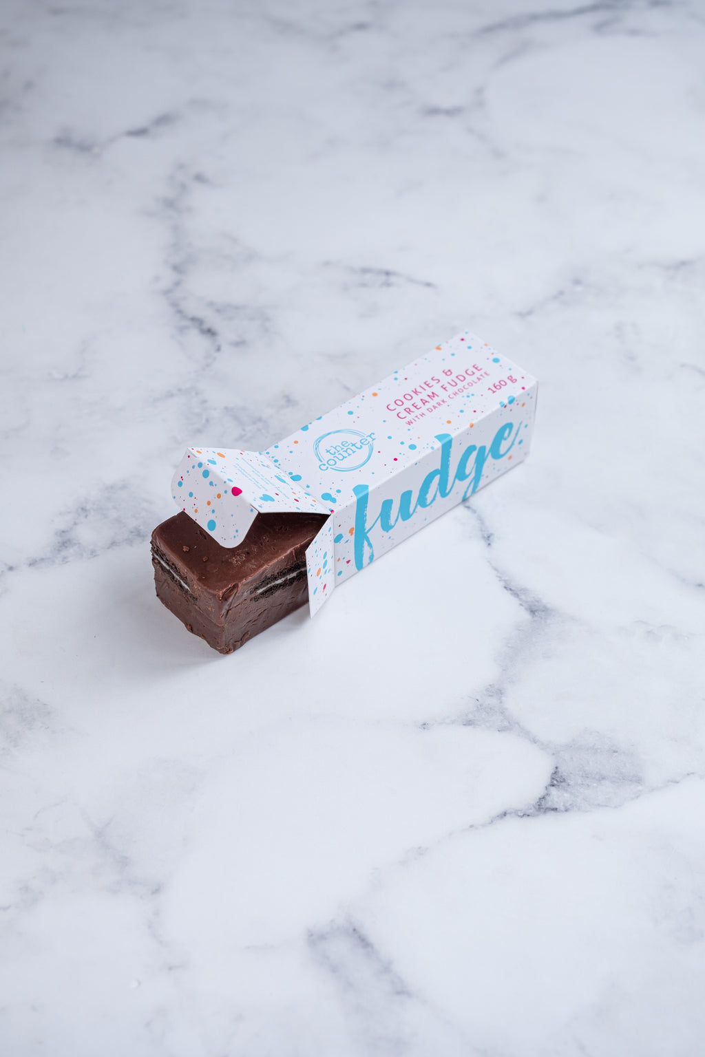 A large bar of dark chocolate and Oreo fudge (cookies and cream) peeks our from a white box. Paint splashes in bright blue and pink surround a messy circled logo for The Counter and the words 'Cookies & Cream fudge with dark chocolate' are written in cerise pink block lettering.