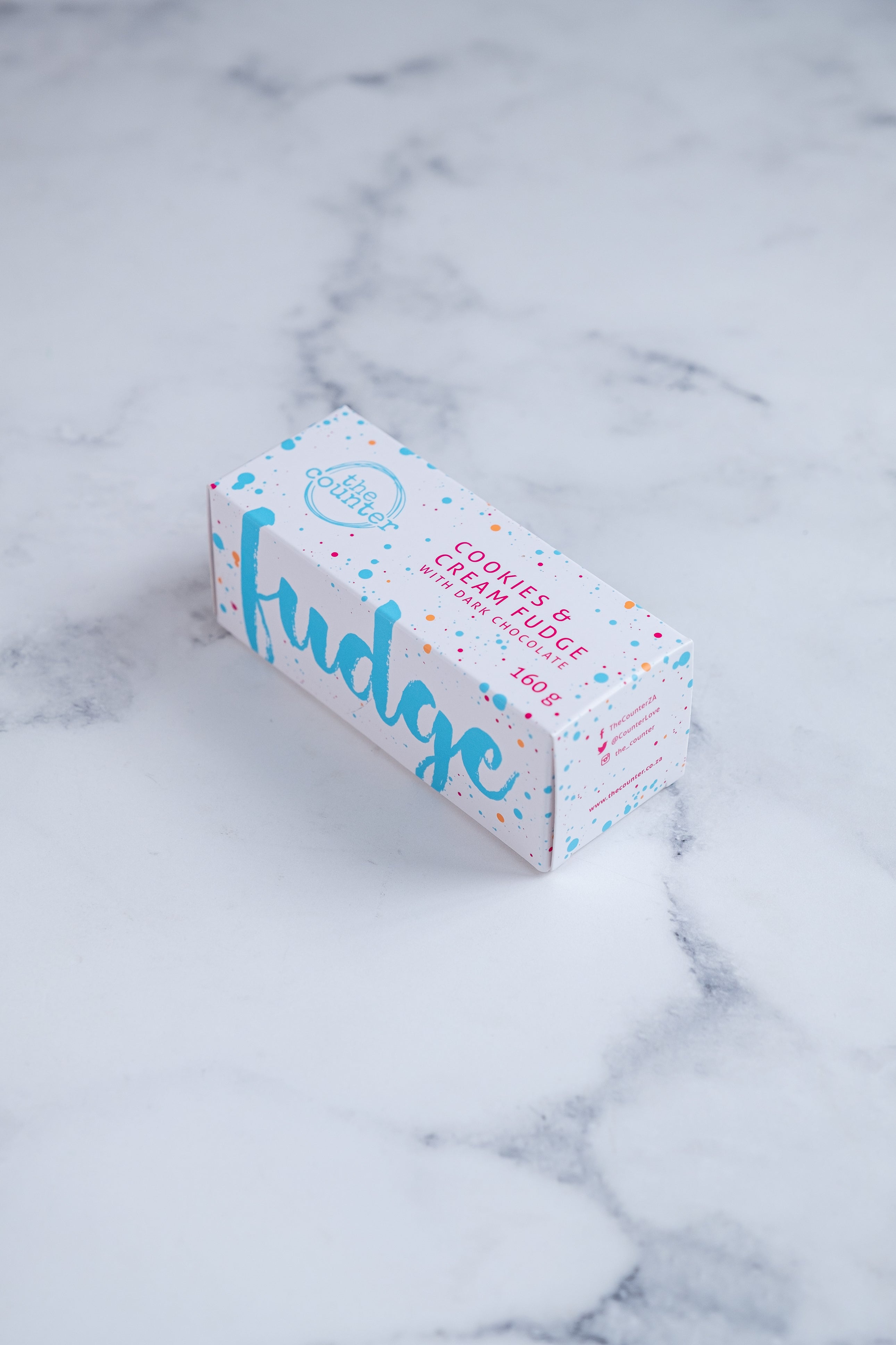 A white box sits on a neutral background. Large bright blue lettering states 'fudge' and is surrounded by bright blue and cerise pink paint splashes. A messy circled logo for The Counter and the words 'Cookies & Cream fudge with dark chocolate' are written in cerise pink block lettering.