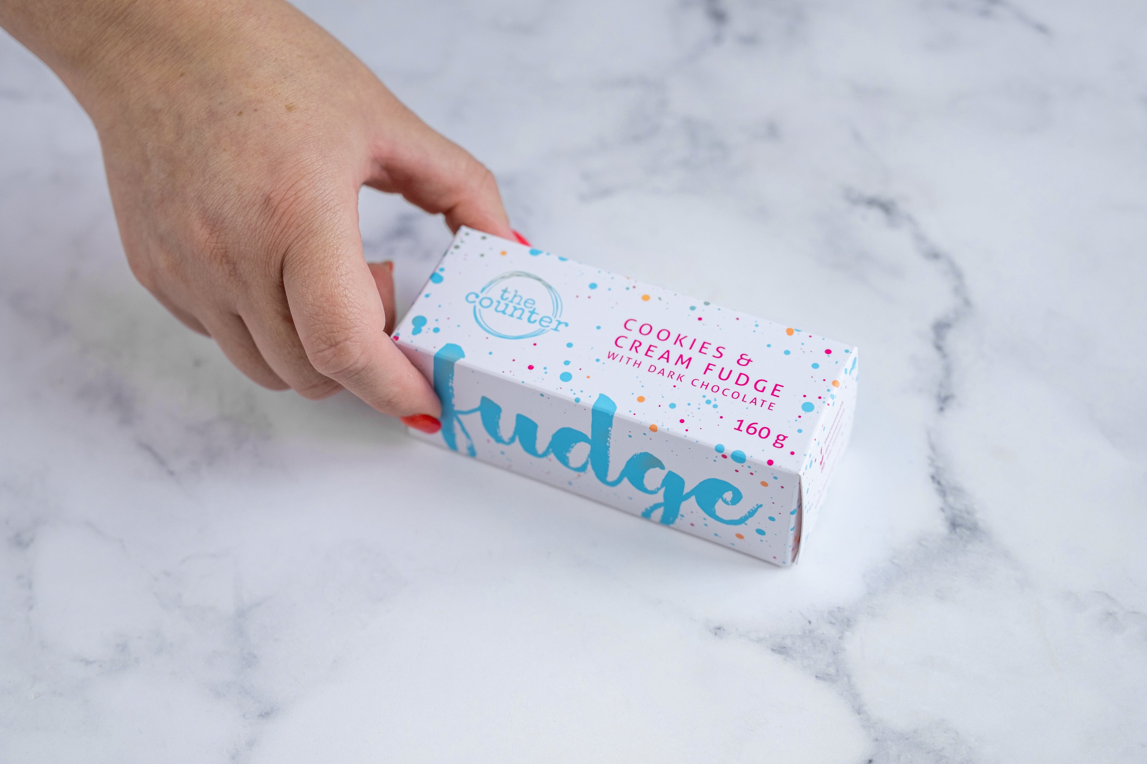 A manicured hand with red nail polish grabs a white box sitting on a neutral background. Large bright blue lettering states 'fudge' and is surrounded by bright blue and cerise pink paint splashes. A messy circled logo for The Counter and the words 'Cookies & Cream fudge with dark chocolate' are written in cerise pink block lettering.