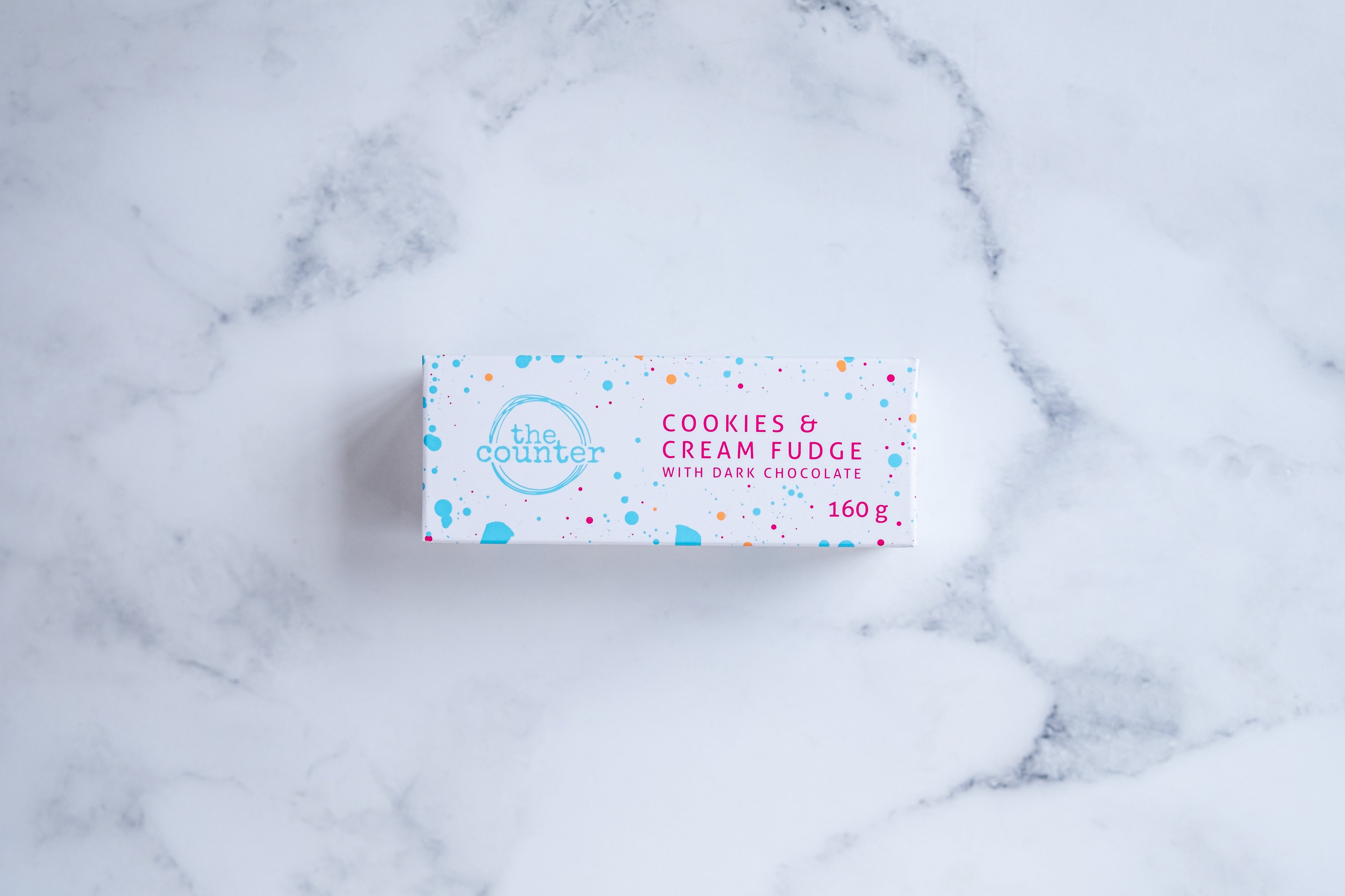 A white box sits on a neutral background. Paint splashes in bright blue and pink surround a messy circled logo for The Counter and the words 'Cookies & Cream fudge with dark chocolate' are written in cerise pink block lettering.