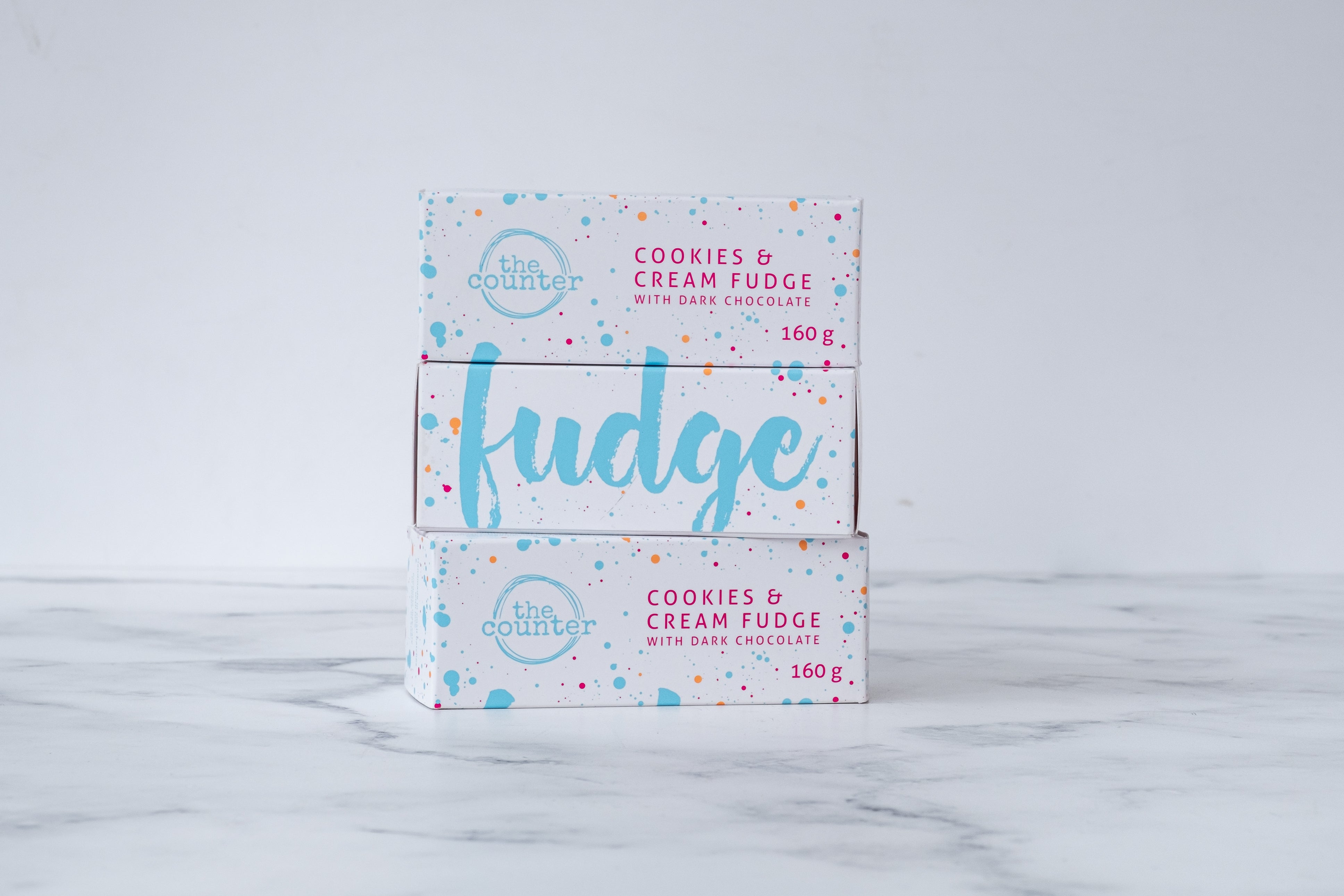 Three white boxes are stacked on a neutral background. Paint splashes in bright blue and pink surround a messy circled logo for The Counter in the first and third box, and the words 'Cookies & Cream fudge with dark chocolate' are written in cerise pink block lettering. The middle box has large, cursive font in bright blue stating the word 'fudge' on it.