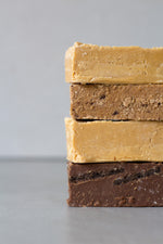 Load image into Gallery viewer, Four large blocks of fudge are stacked on top of each other, peeking out from the right side of the image - each in a different flavour and colour. The second block from the top is a slightly brown Coco Malt fudge bar.
