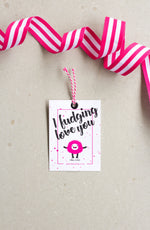 Load image into Gallery viewer, Cerise pink and white striped ribbon is twisted and is sits on a neutral surface - underneath this ribbon is a white swing tag, with a very light cerise pink border about 1cm in. Large black text says &#39;I fudging love you&#39; above a cute pink and black monster icon. The swing tag is dotted with subtle pink splashes of oclour, and is finished with pink and white twine, enrobed through a black eyelet.
