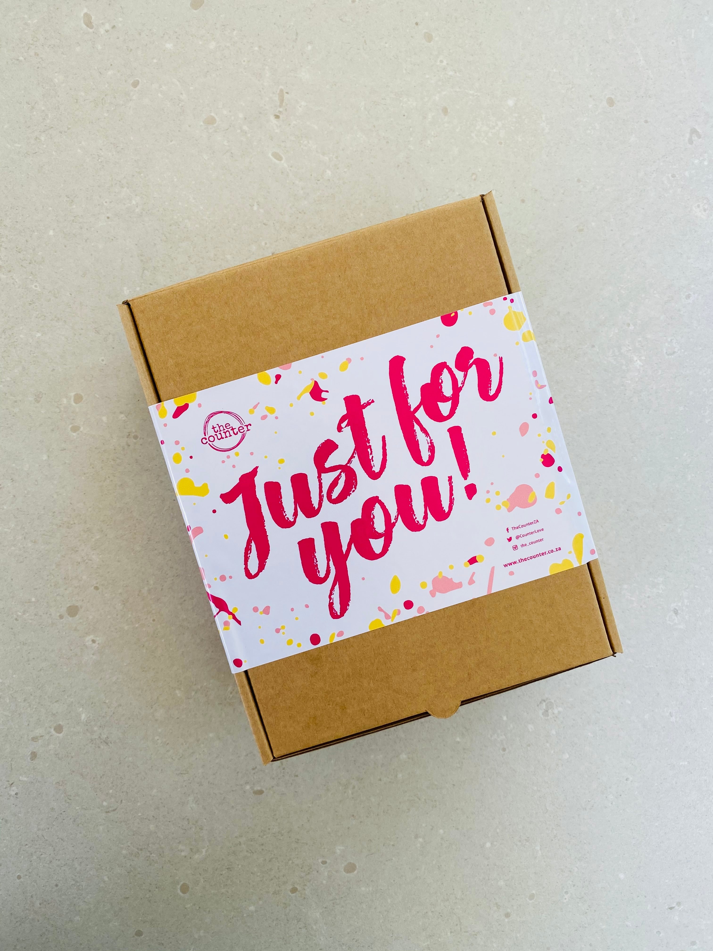 A brown gift box sits on a neutral surface. It is finished with a large, brightly coloured sticker with the text 'Just for You!' written in cerise pink. The sticker is decorated with brightly coloured paint splashes in cerise, light pink and yellow - with The Counter' logo in the top left of the sticker.