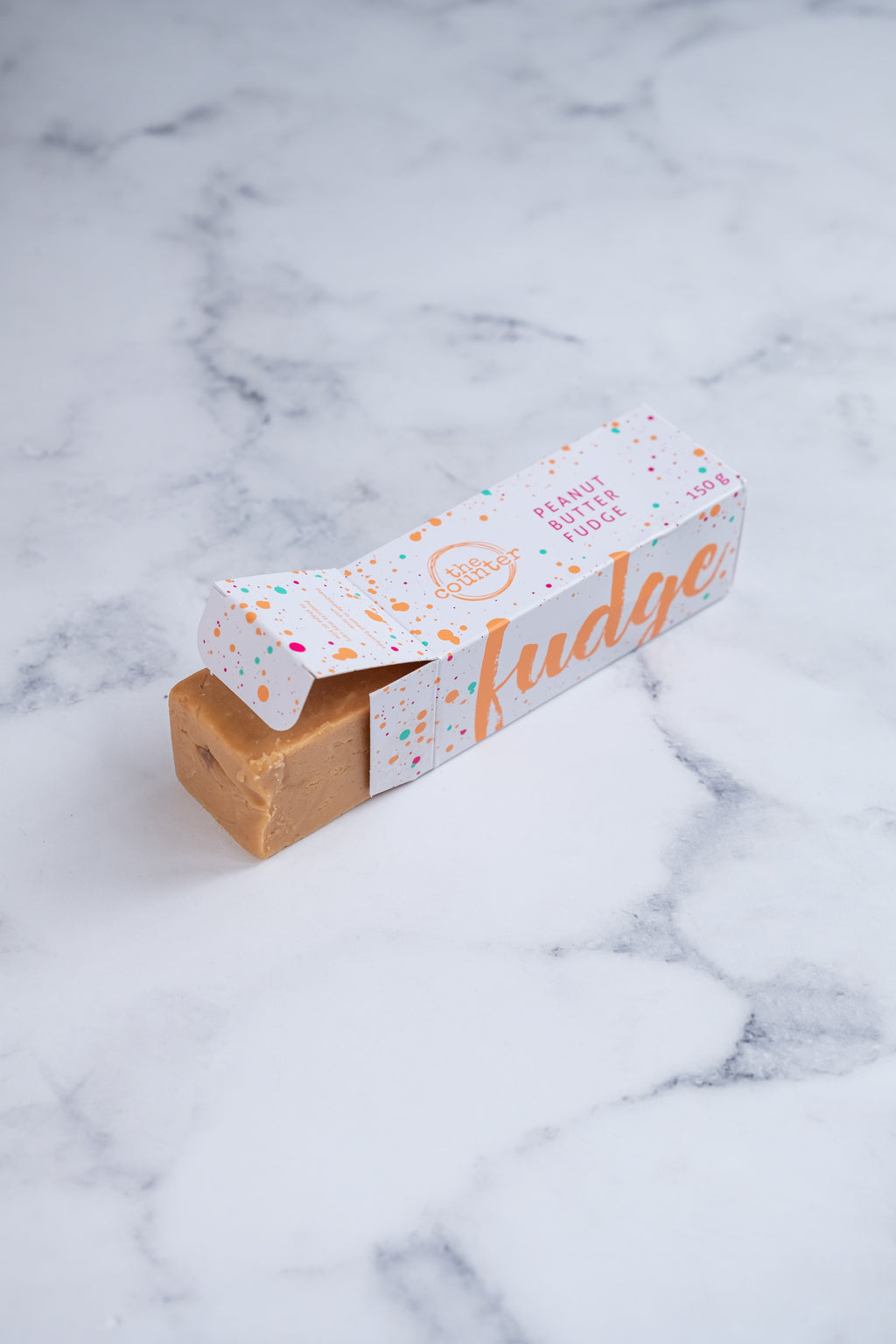 A large bar of peanut butter fudge peeks our from a white box. Paint splashes in bright orange and pink surround a messy circled logo for The Counter and the words 'Peanut Butter fudge' are written in cerise pink block lettering.