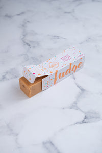 A large bar of peanut butter fudge peeks our from a white box. Paint splashes in bright orange and pink surround a messy circled logo for The Counter and the words 'Peanut Butter fudge' are written in cerise pink block lettering.