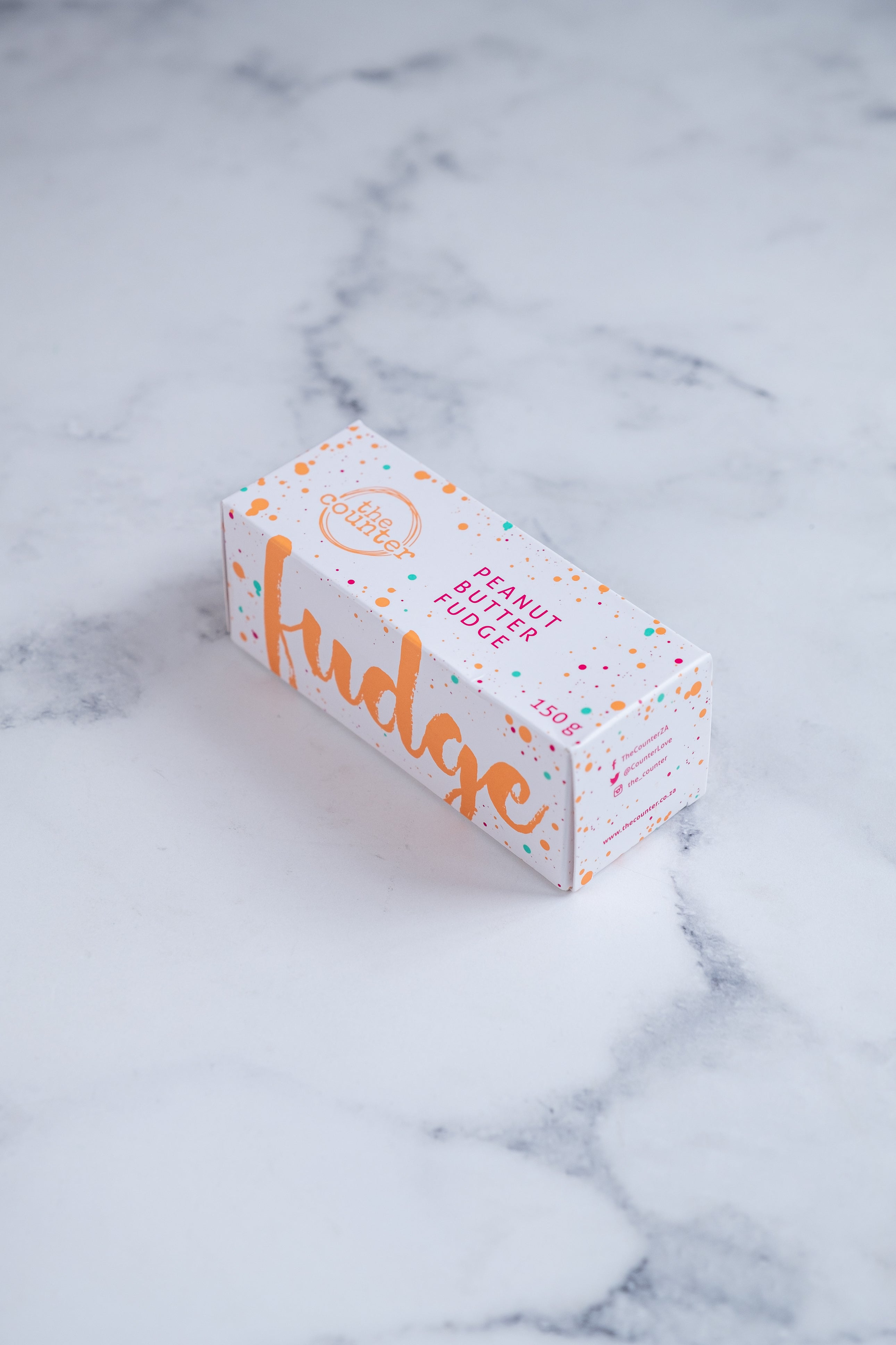 A white box sits on a neutral background. Paint splashes in orange and pink surround a messy circled logo for The Counter and the words ‘Peanut Butter Fudge' and 'Fudge' are written on different sides of the box in cerise pink block lettering.