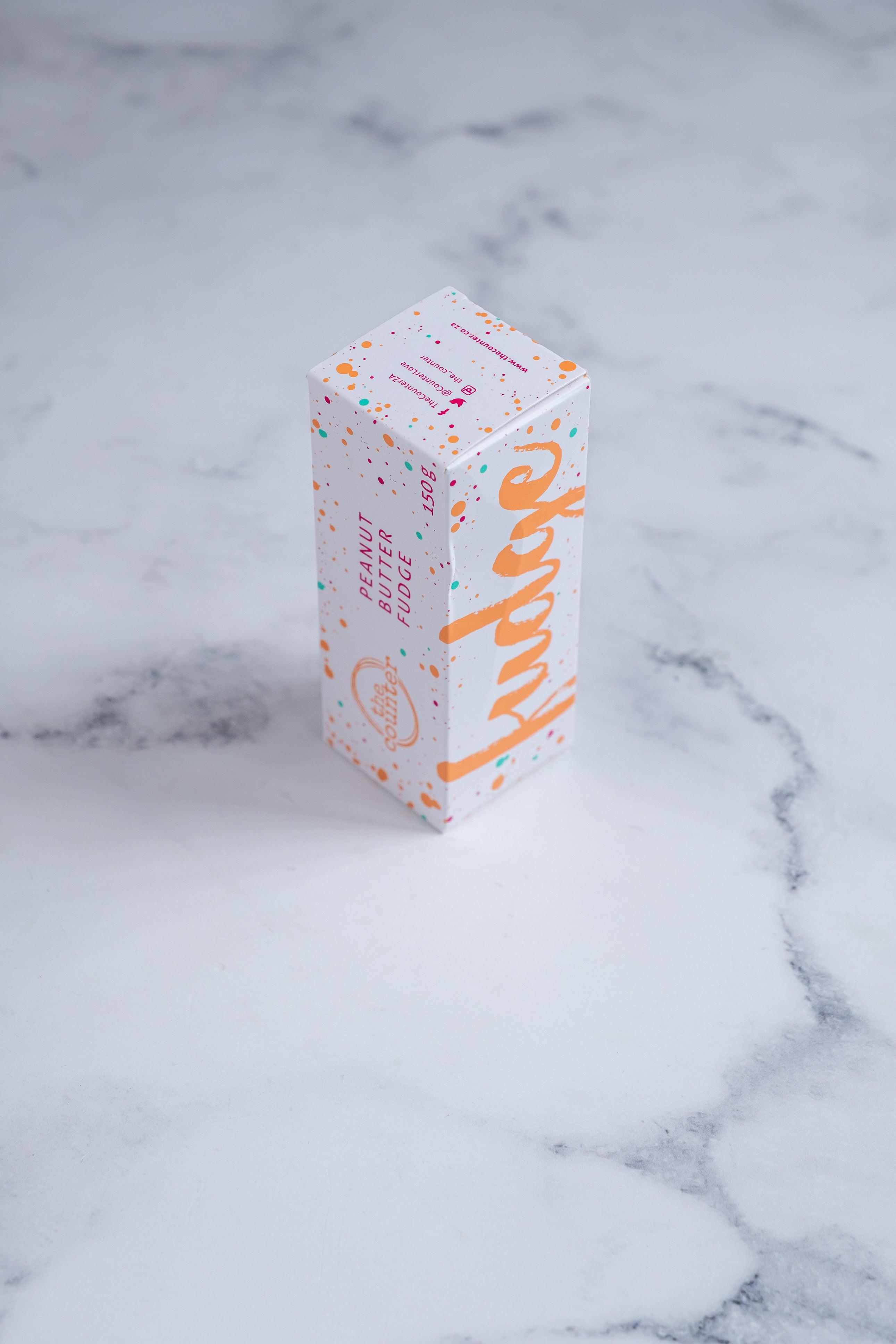 A white box sits on a neutral background. Paint splashes in orange and pink surround a messy circled logo for The Counter and the words ‘Peanut Butter Fudge' and 'Fudge' are written on different sides of the box in cerise pink block lettering.