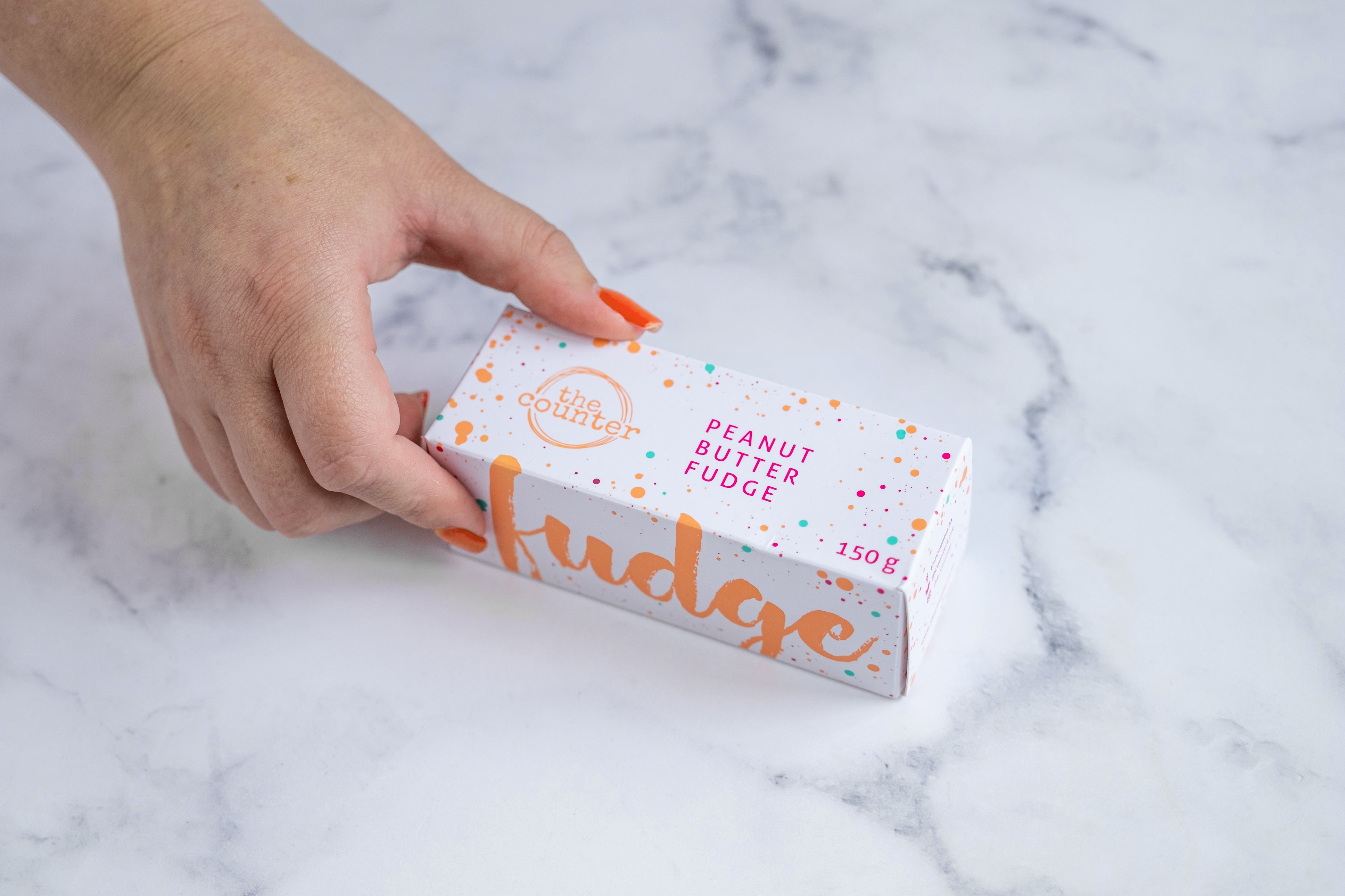 A white box sits on a neutral background. Paint splashes in orange and pink surround a messy circled logo for The Counter and the words ‘Peanut Butter Fudge' and 'Fudge' are written in cerise pink block lettering and cursive orange lettering respectively, on either side of the box. A manicured hand with red nail polish grabs the box.