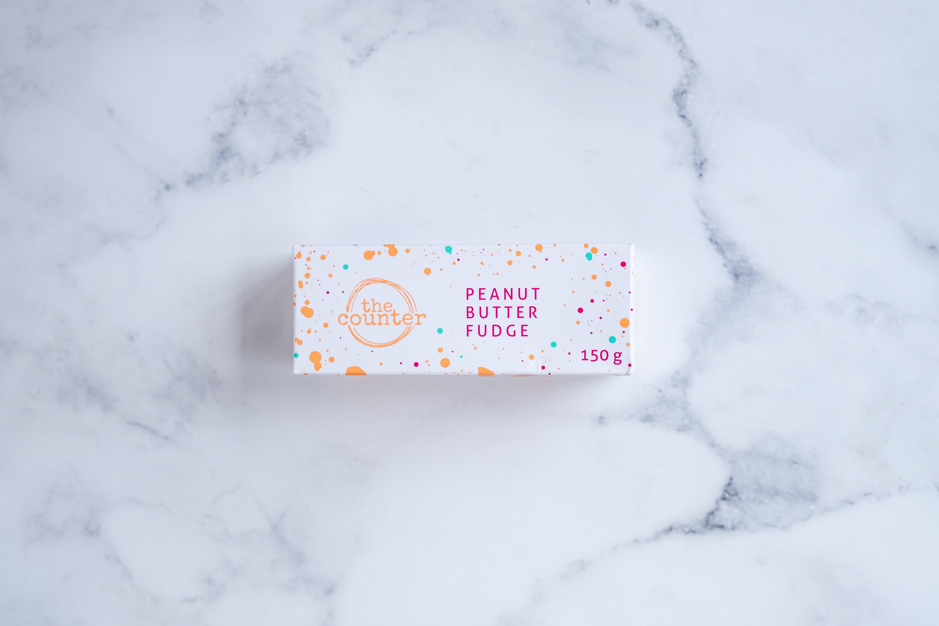 A white box sits on a neutral background. Paint splashes in orange and pink surround a messy circled logo for The Counter and the words ‘Peanut Butter Fudge' are written in cerise pink block lettering.