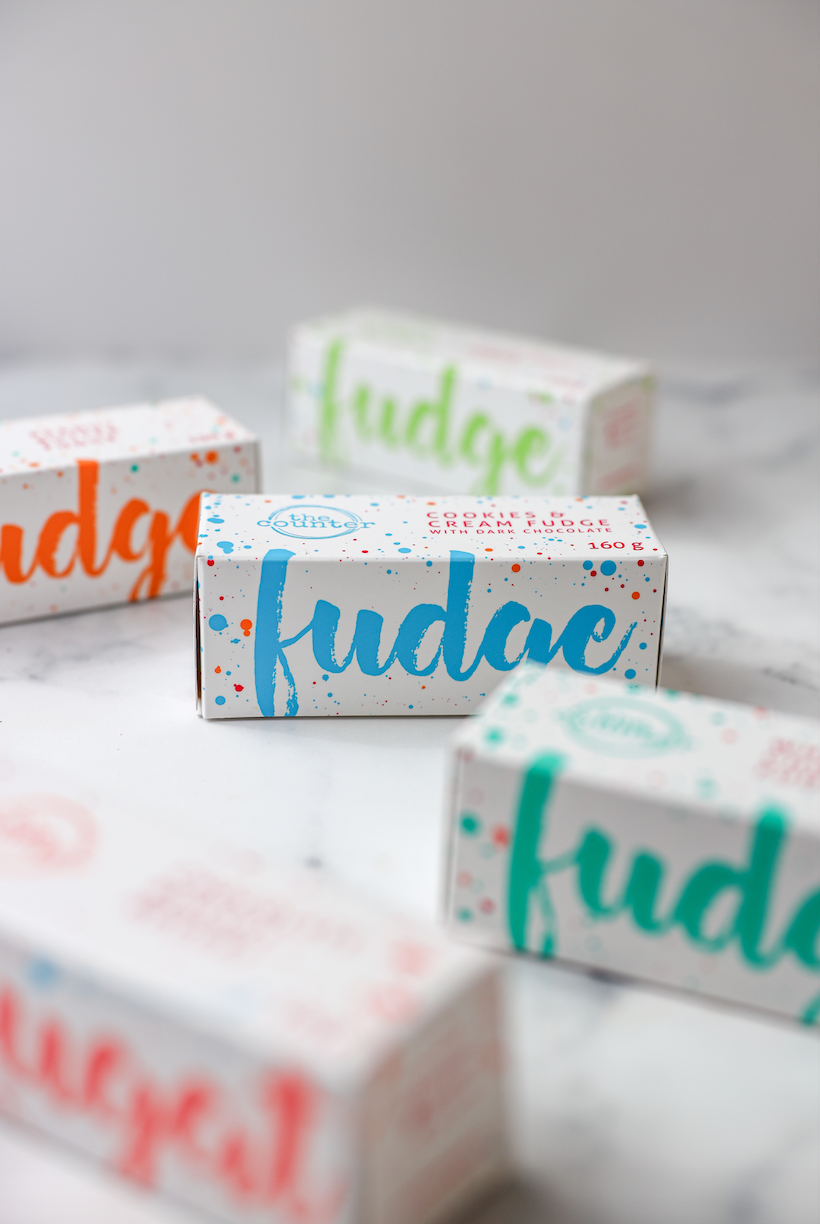 Five colourful fudge boxes sit on a marbled white surface, with the Cookies and Cream fudge box being the only one in focus. The box is white with bright blue large text stating 'fudge' and a subtle logo and product description on the top of the box.