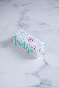 White fudge box with large, cursive turquoise text and colourful paint splashes around it; sitting on a white and grey marbled surface.
