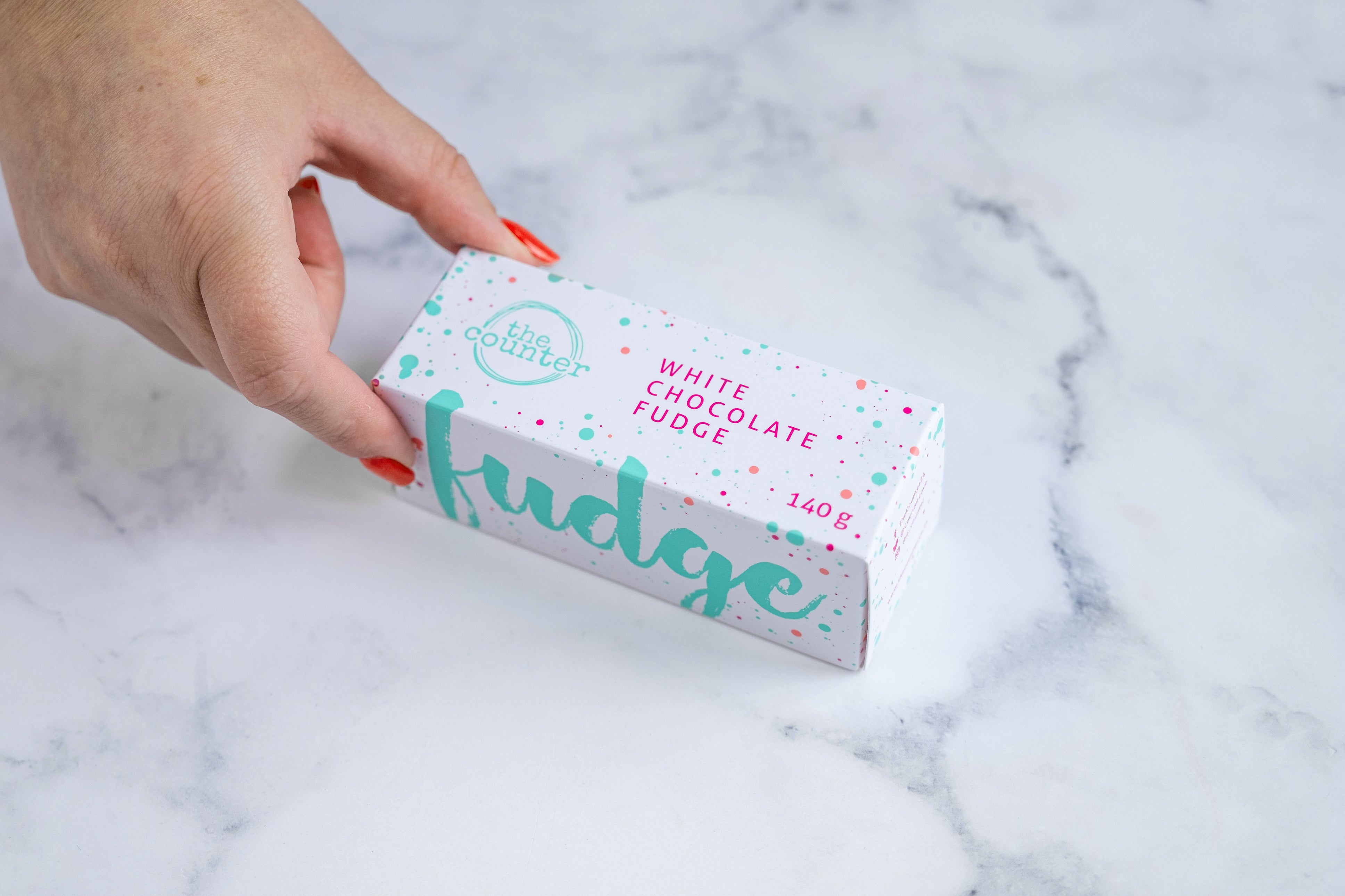 A hand with red nail polish grabs a White chocolate fudge box with 'The Counter' logo in turquoise, surrounded by several thin lines creating a circle. Pink text stating 'white chocolate fudge' and paint splashes surrounding the text and logo.