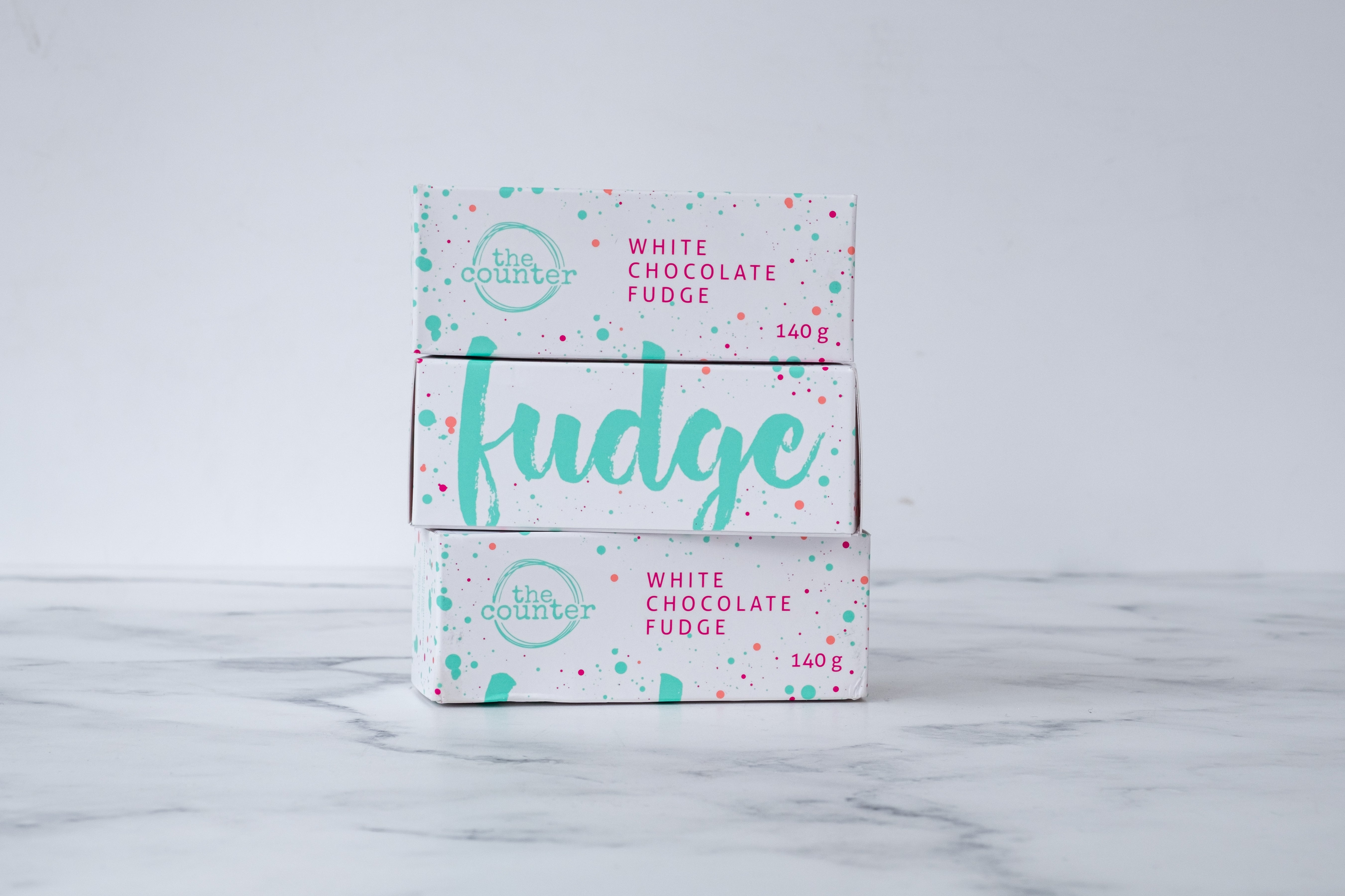 Three white fudge boxes with large, cursive turquoise text and colourful paint splashes around it; sitting on a white and grey marbled surface. Three boxes are stacked on top of each other; alternating between different sides of the box.